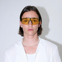 A person with straight brown hair, wearing Fast Lane Sunglasses in black and acid with UV400 protection, along with a white shirt and a pearl necklace, stands against a plain white background.