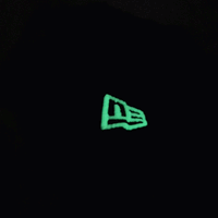 A glowing New Era logo stands out against a deep black backdrop, lit up in green while the surrounding image stays dark—mirroring the unique aesthetic of the chainstitch embroidery found on the San Francisco Giants All-Star Trolley Corduroy 19Twenty Snapback Hat in black and orange.