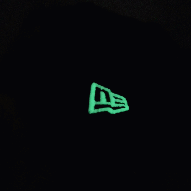 A glowing New Era logo stands out against a deep black backdrop, lit up in green while the surrounding image stays dark—mirroring the unique aesthetic of the chainstitch embroidery found on the San Francisco Giants All-Star Trolley Corduroy 19Twenty Snapback Hat in black and orange.