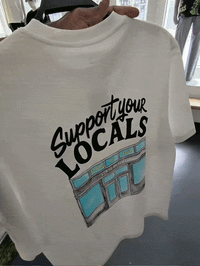 The Support Your Locals Tee in white features a detailed storefront graphic with the bold phrase “Support your LOCALS” in black lettering across the back and a “STAY OPEN” store sign, symbolizing community resilience. Held up indoors, this standout tee is part of State Of Flux San Francisco’s collection.
