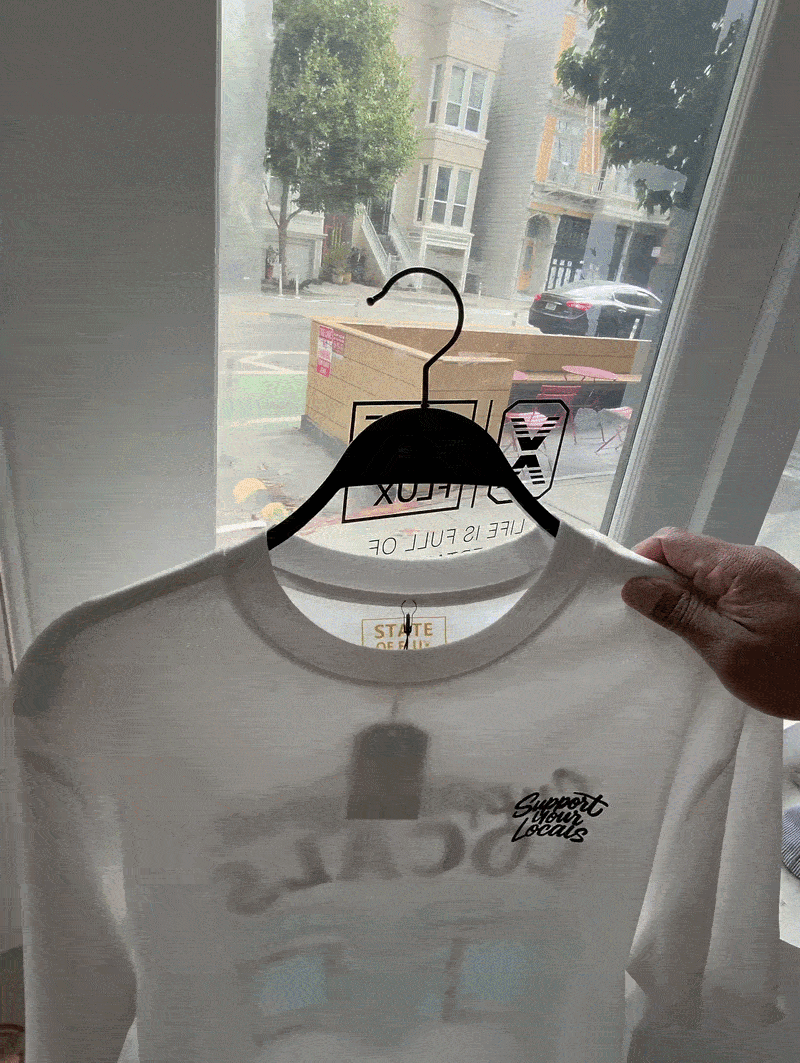 A hand holds up a white Support Your Locals Tee inside the State Of Flux San Francisco storefront, with boxes and buildings visible outside through large windows.