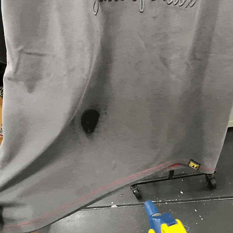 A heat gun is aimed at the SOF Watermark Script Tee in gradient grey, shrinking a wet spot and drying the fabric while the water-reactive tee hangs on a rack.