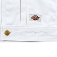 Close-up of the Selvedge Pleated Denim Jacket in white, featuring a gold button and logo patch sewn near the hem. The diagonal weave adds classic style and durability to this workwear-inspired piece.