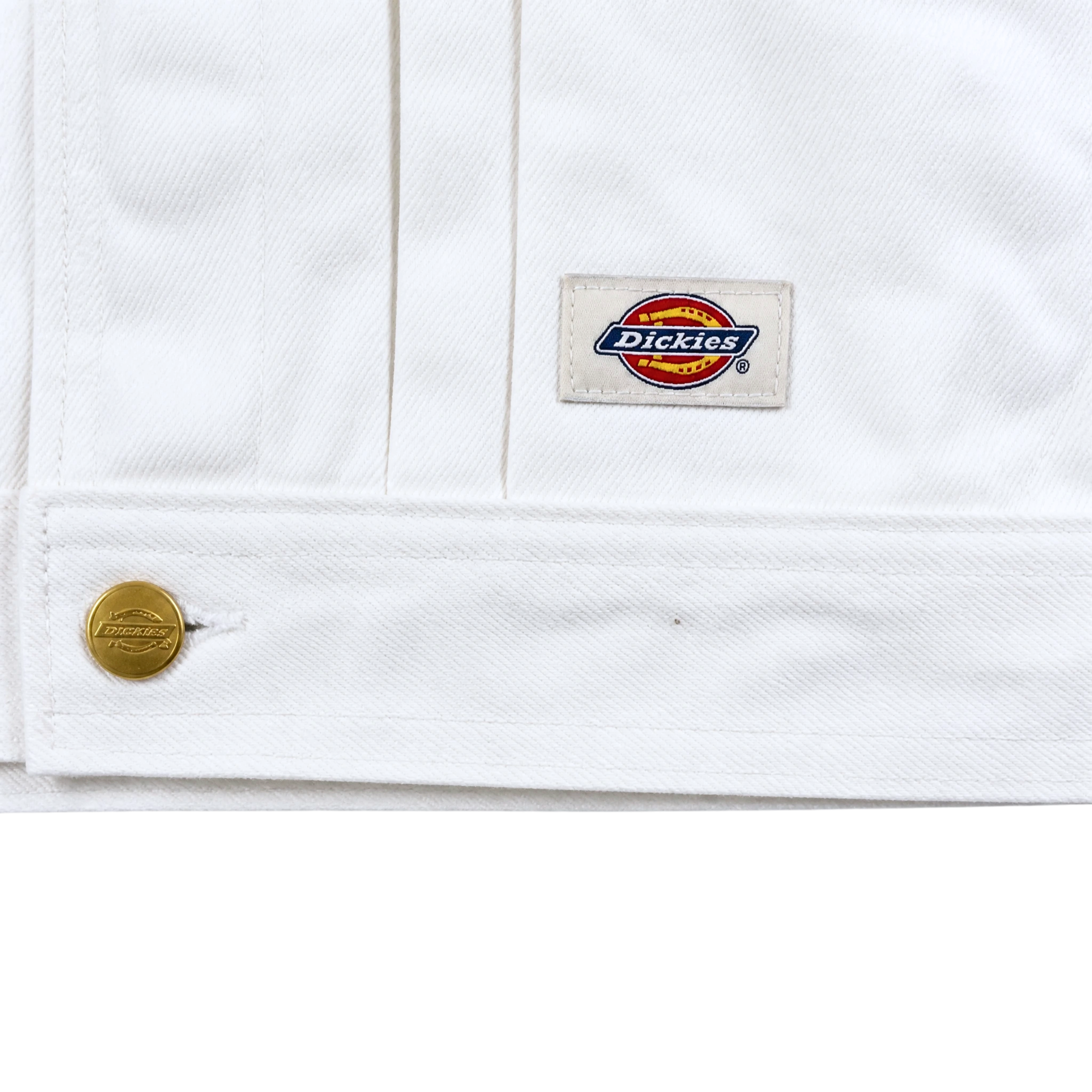 Close-up of the Selvedge Pleated Denim Jacket in white, featuring a gold button and logo patch sewn near the hem. The diagonal weave adds classic style and durability to this workwear-inspired piece.