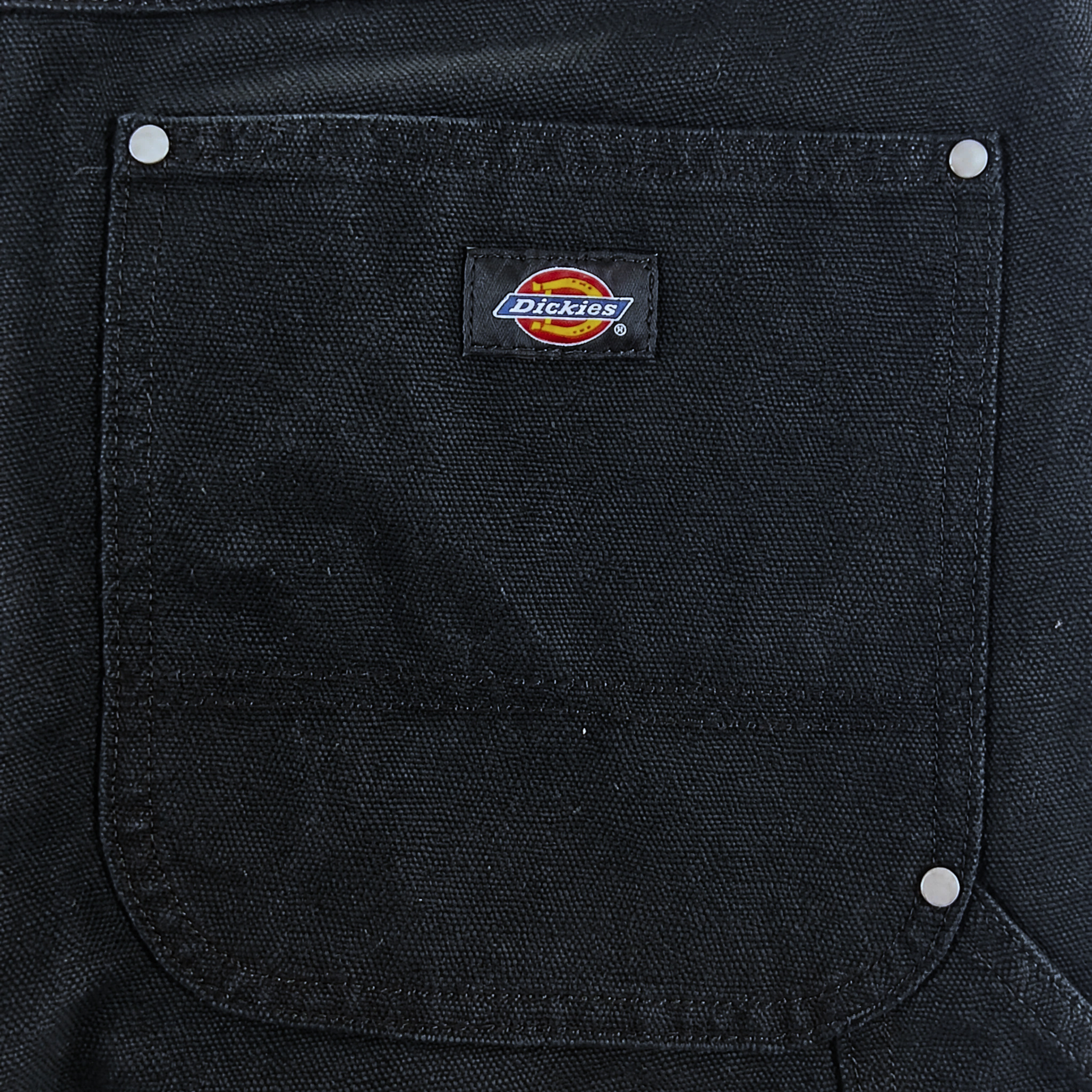 Close-up of a black fabric pocket with silver rivets and a sewn-on Dickies logo patch, showcasing the durable construction of the Double Knee Canvas Shorts in black.