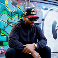 A man in a black hoodie, sunglasses, and a PREORDER ONLY San Francisco 49ers Golden Ticket V2 59Fifty Fitted Hat in black and cardinal sits before a vibrant graffiti wall, elbows on his knees, looking down.