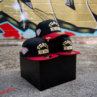 Three black and cardinal San Francisco 49ers Golden Ticket V2 59Fifty Fitted Hats (PREORDER ONLY) are displayed on a black cube outdoors, with a graffiti-covered wall in the background.