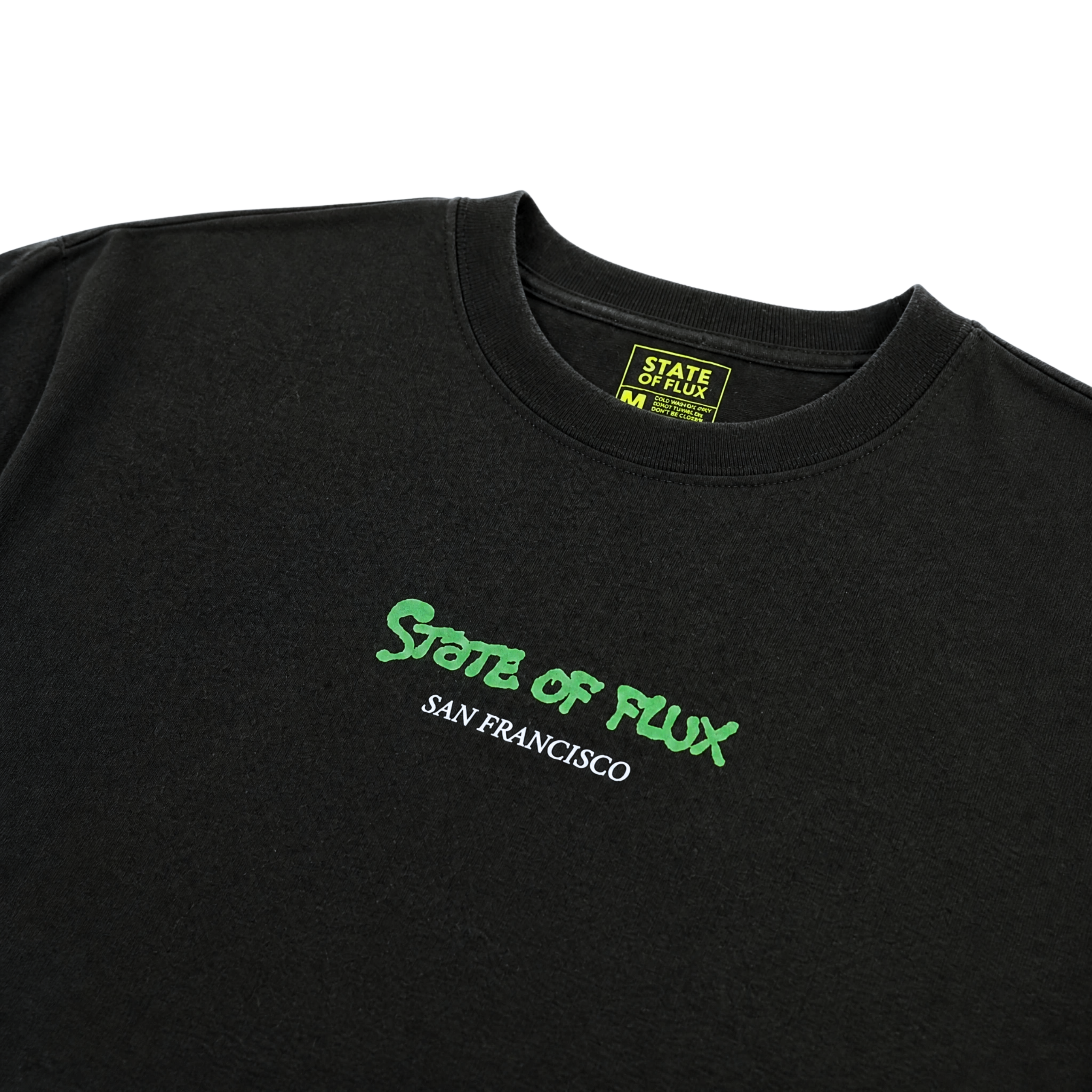The Keep Goin' Tee in black is a heavyweight cotton, oversized streetwear t-shirt with STATE OF FLUX in green and SAN FRANCISCO in white on the chest, inspired by SF's Mission District. Features a yellow & black collar tag with the brand name and size.