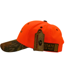 The Peep Game Hat in safety orange and mossy oak features a camouflage-patterned brim, “STATE OF FLUX” side patch, and wooden Mossy Oak tag—ideal high-visibility headwear for the outdoors.