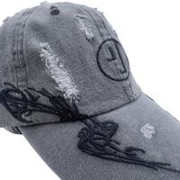 A close-up of the Everyday Is A Gift Cap in washed black—a gray, distressed denim baseball hat featuring torn patches, black embroidered details, and a circular logo on the front—an effortlessly cool accessory perfect for streetwear looks.
