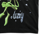A green alien with clasped hands sits above the word “Anxiety” on a black fabric background, surrounded by white sparkling stars, creating a bold streetwear aesthetic. This graphic appears on the Anxiety Flower Tee in black, crafted from heavyweight cotton.
