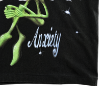 A green alien with clasped hands sits above the word “Anxiety” on a black fabric background, surrounded by white sparkling stars, creating a bold streetwear aesthetic. This graphic appears on the Anxiety Flower Tee in black, crafted from heavyweight cotton.