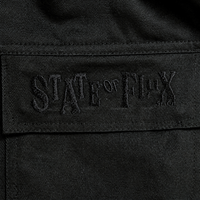 Close-up of black fabric on the Flux Utility Cargo Pants in black, featuring "STATE OF FLUX" embroidered in matching black thread for a subtle, textured effect. The embroidery appears on a rectangular patch stitched onto the pants, echoing the signature detailing found on premium cargo pants with utility pockets.