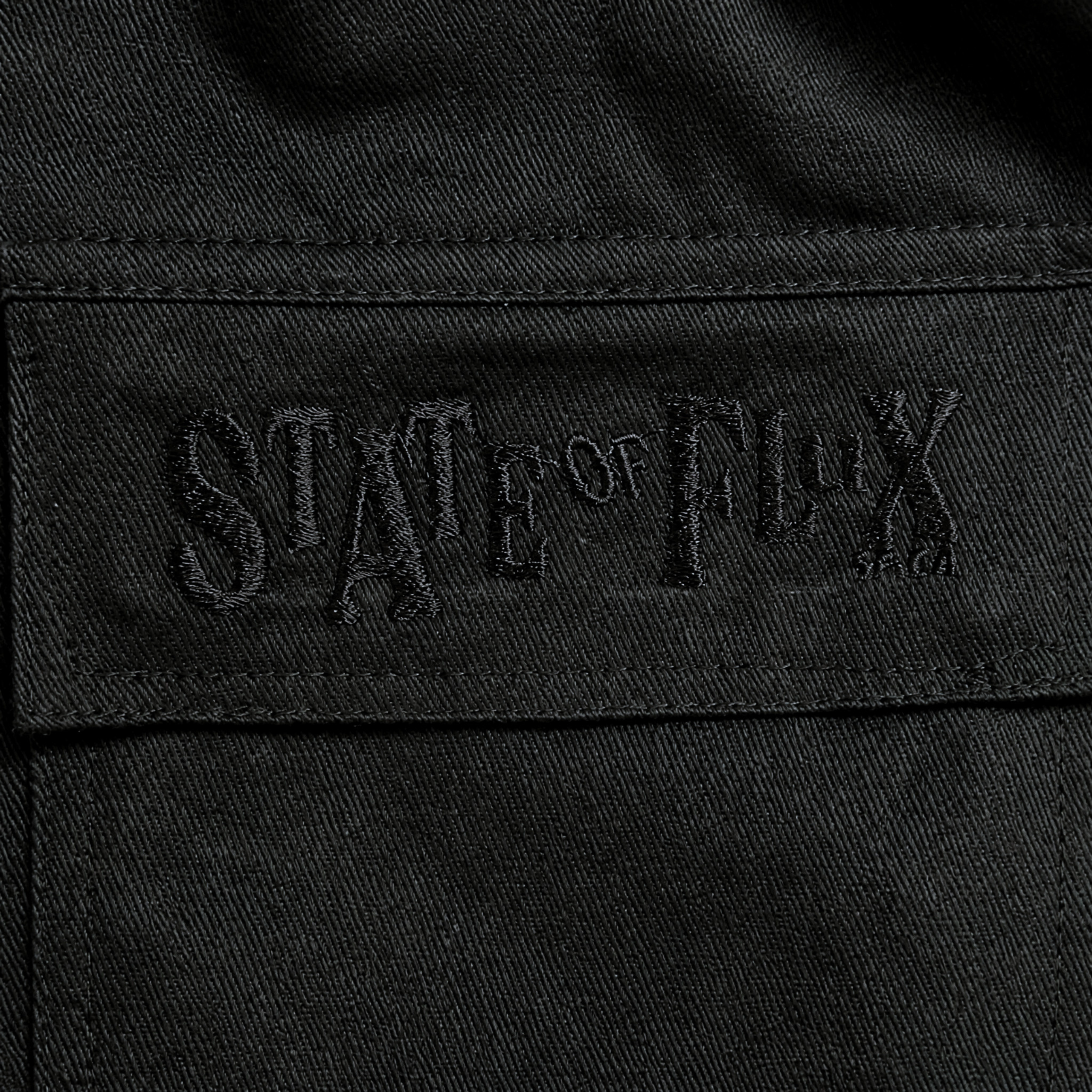 Close-up of black fabric on the Flux Utility Cargo Pants in black, featuring "STATE OF FLUX" embroidered in matching black thread for a subtle, textured effect. The embroidery appears on a rectangular patch stitched onto the pants, echoing the signature detailing found on premium cargo pants with utility pockets.