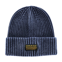 The Ribbed Knit Mantra Beanie in washed navy features a dark ribbed knit design with a folded brim and a yellow patch that reads STATE OF FLUX alongside an inspiring quote. Made from a comfortable rayon blend, this cozy accessory merges style and motivation in one versatile beanie.
