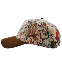Introducing the Wildflower Strapback Hat in cream and brown: this strapback features a brown suede brim and a floral tapestry pattern on the crown adorned with intricate botanical artwork. A small black tag on the side reads STATE OF FLUX.