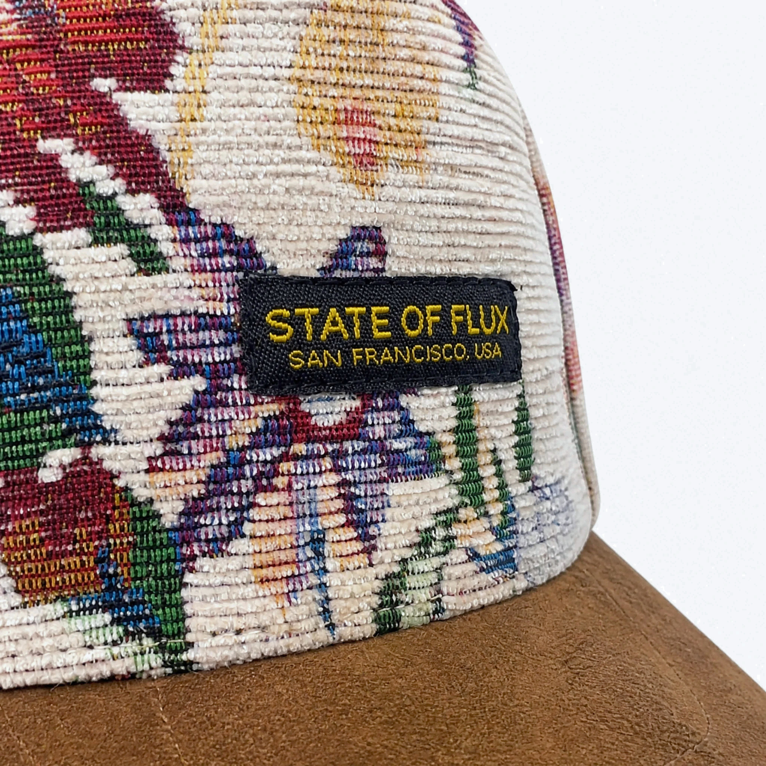 The Wildflower Strapback Hat in cream and brown features a floral jacquard woven pattern and a brown brim. A black tag on the front displays “STATE OF FLUX SAN FRANCISCO, USA” in yellow text, accentuating its unique botanical artwork for a stylish look.