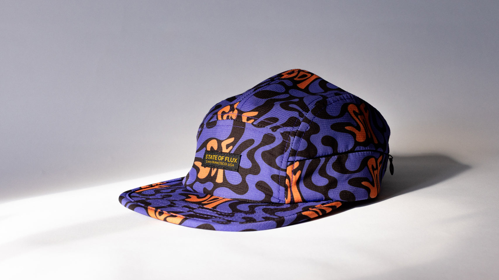 A purple 5-panel cap with an orange and black abstract wavy pattern, featuring a small black and yellow 'STATE OF FLUX' brand patch on the front, set against a gray studio background with a strong diagonal shadow.